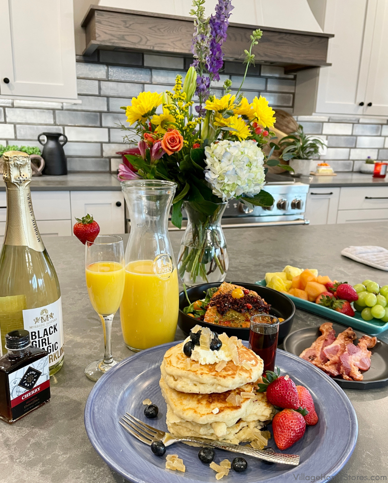 island countertop full of brunch food including stack of pancakes on blue plate and mimosas